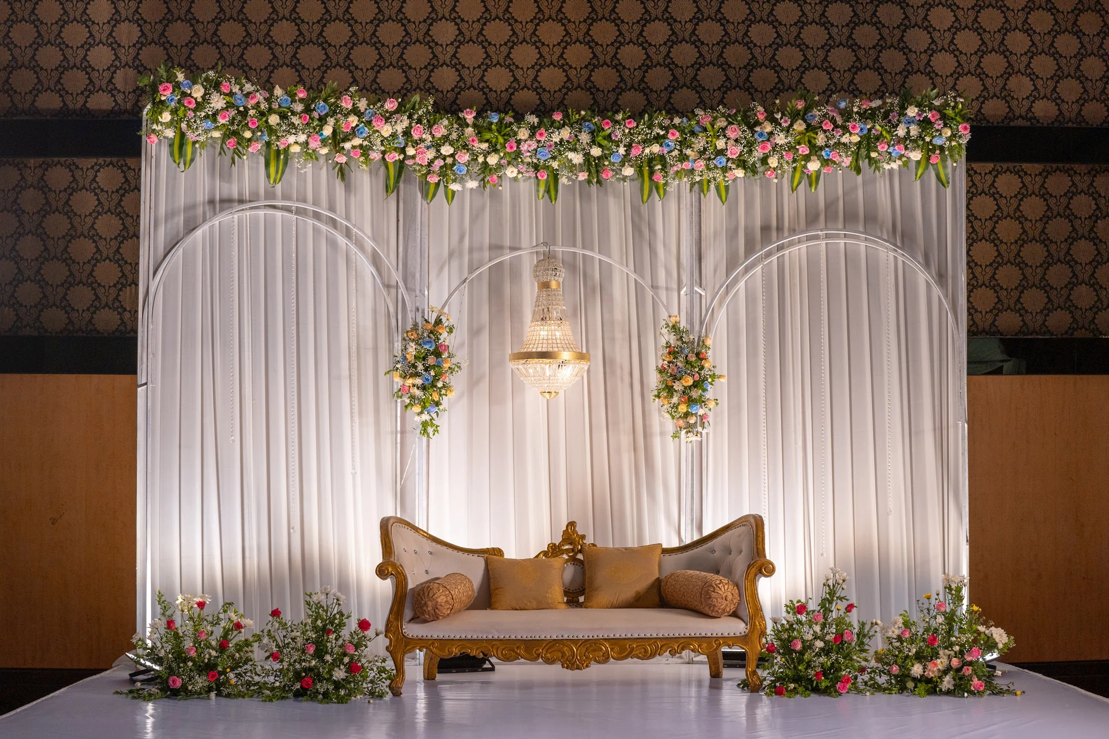 An indoor celebration stage arranged with ivory drapes, flowers, a chandelier and guest seating