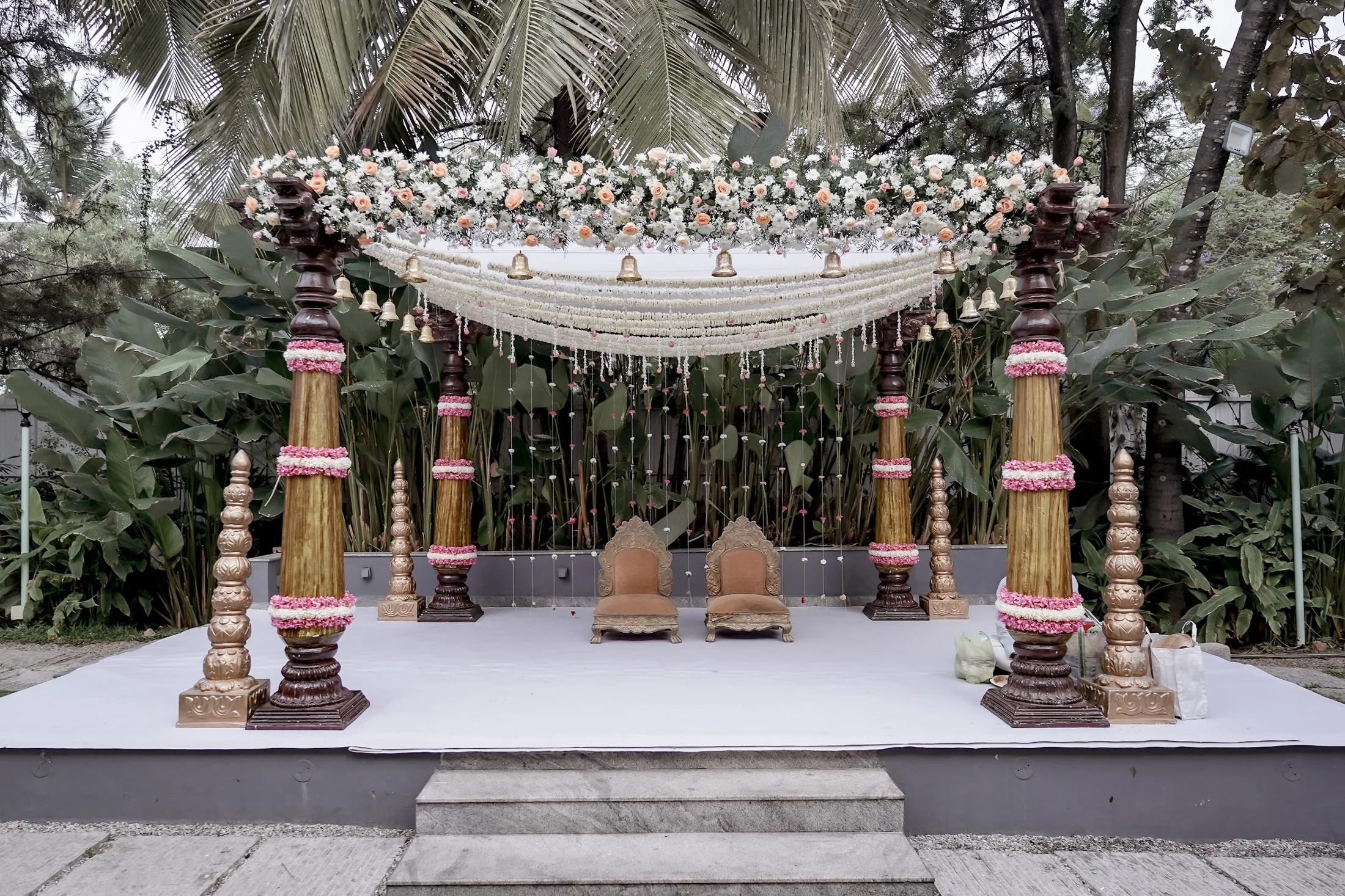 Carved pillars, white flower garlands and brass bells framing an outdoor ceremony mandap.