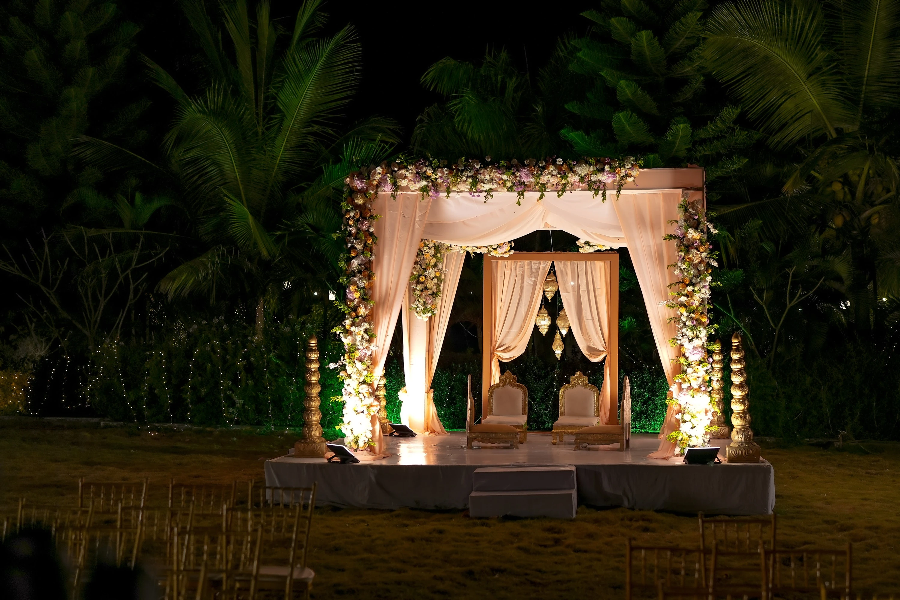 A warmly lit mandap and flower-framed drapes in an evening garden