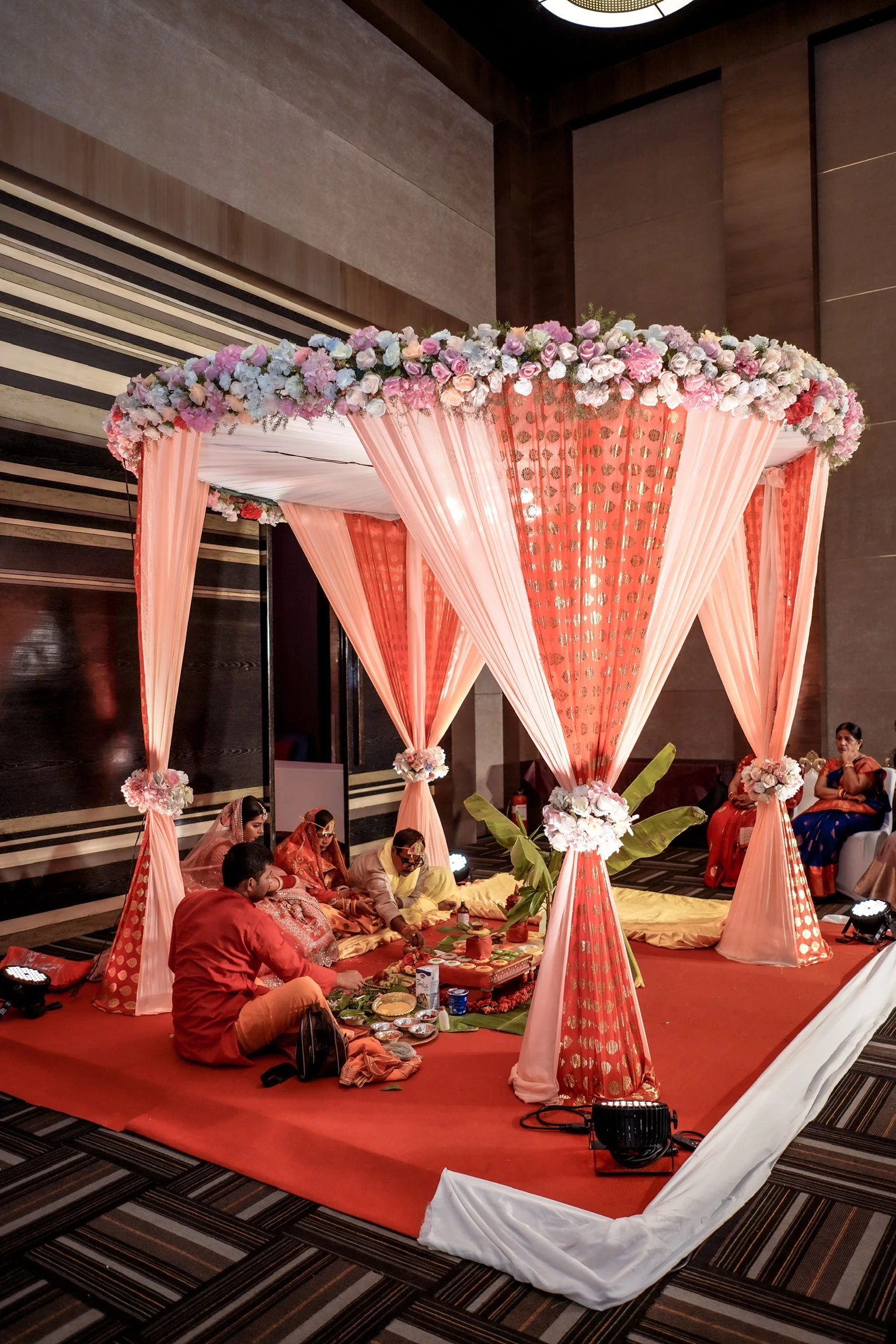 A pink indoor mandap framed with flowers and brass ceremony vessels