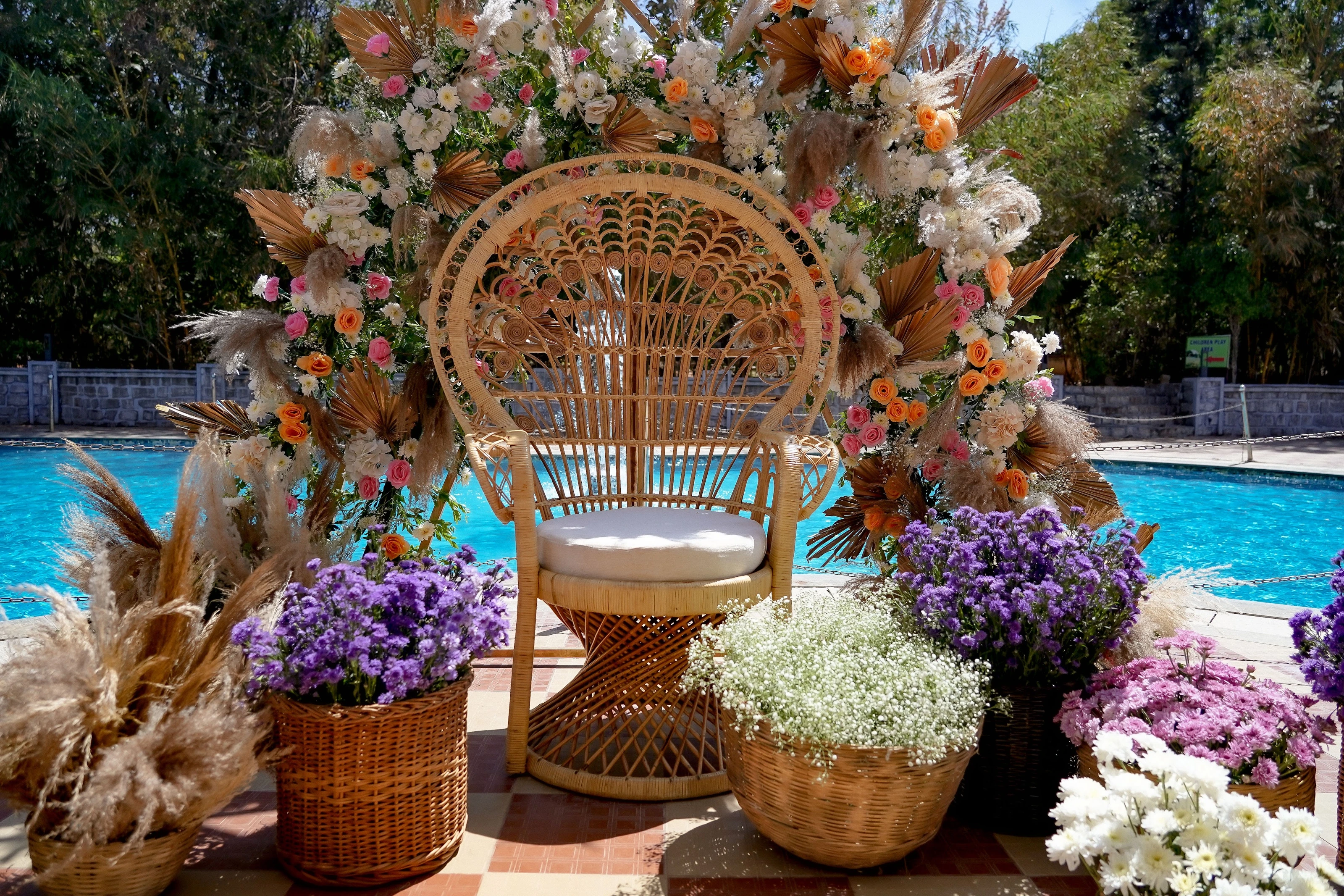 A wicker mehendi seat surrounded by pastel flowers and baskets beside a pool