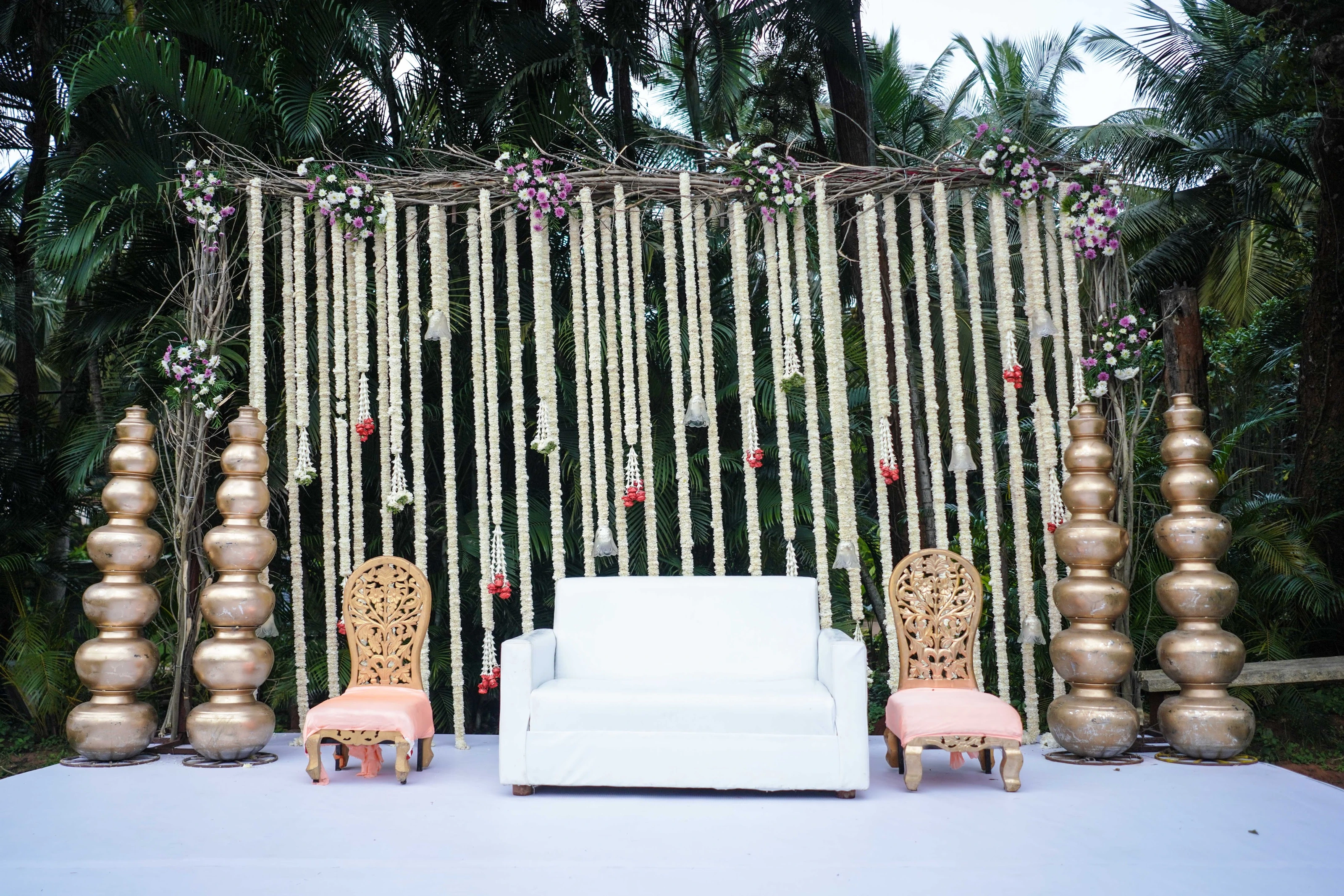 A white sofa and carved chairs beneath hanging flower garlands and brass accents