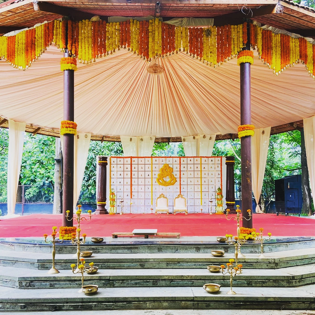 A covered ceremony pavilion with orange and yellow garlands and brass lamps